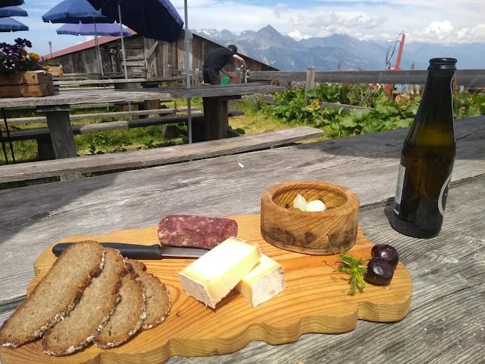 Wooden board with bread, cheese, sausage, and a bottle on an outdoor table in the Alps at Bovine