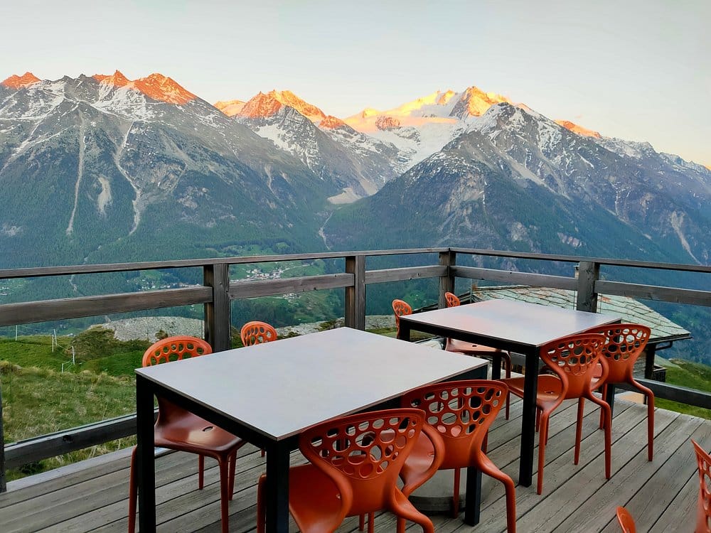 Outdoor restaurant terrace with tables and orange chairs overlooking snow-capped alpine mountains at sunset