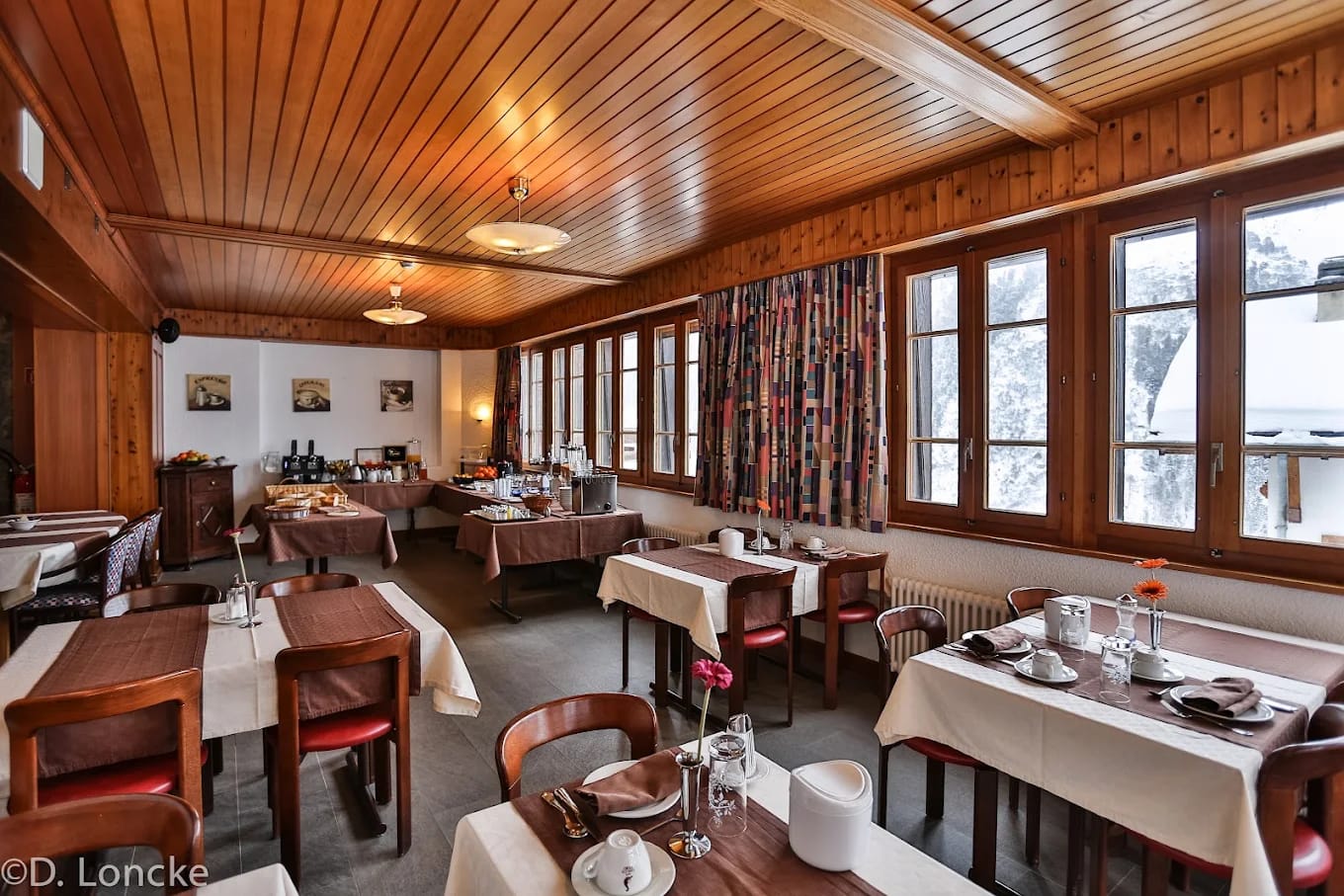 Hotel dining room with wooden panel ceiling, tables set with white tablecloths, and large windows overlooking snow