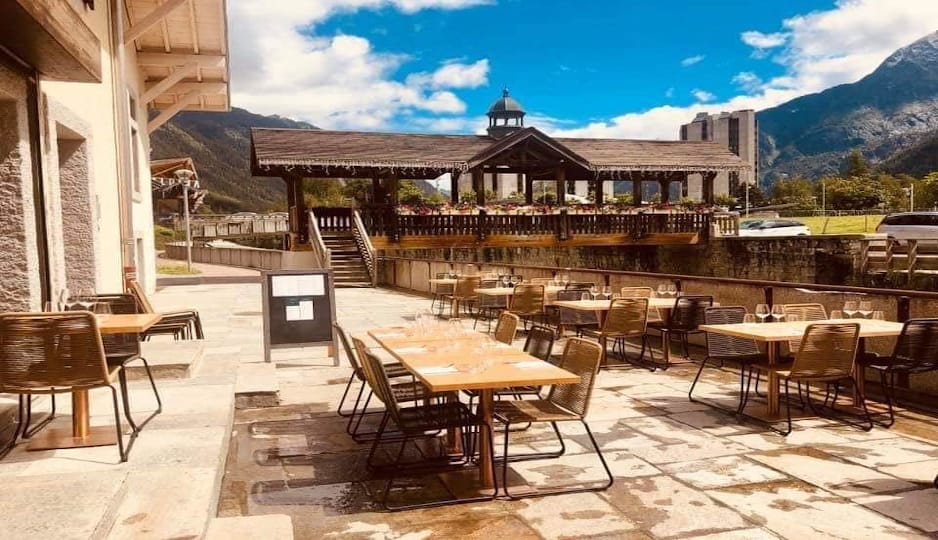 Outdoor cafe terrace with wooden tables and chairs facing a mountain stream and bridge