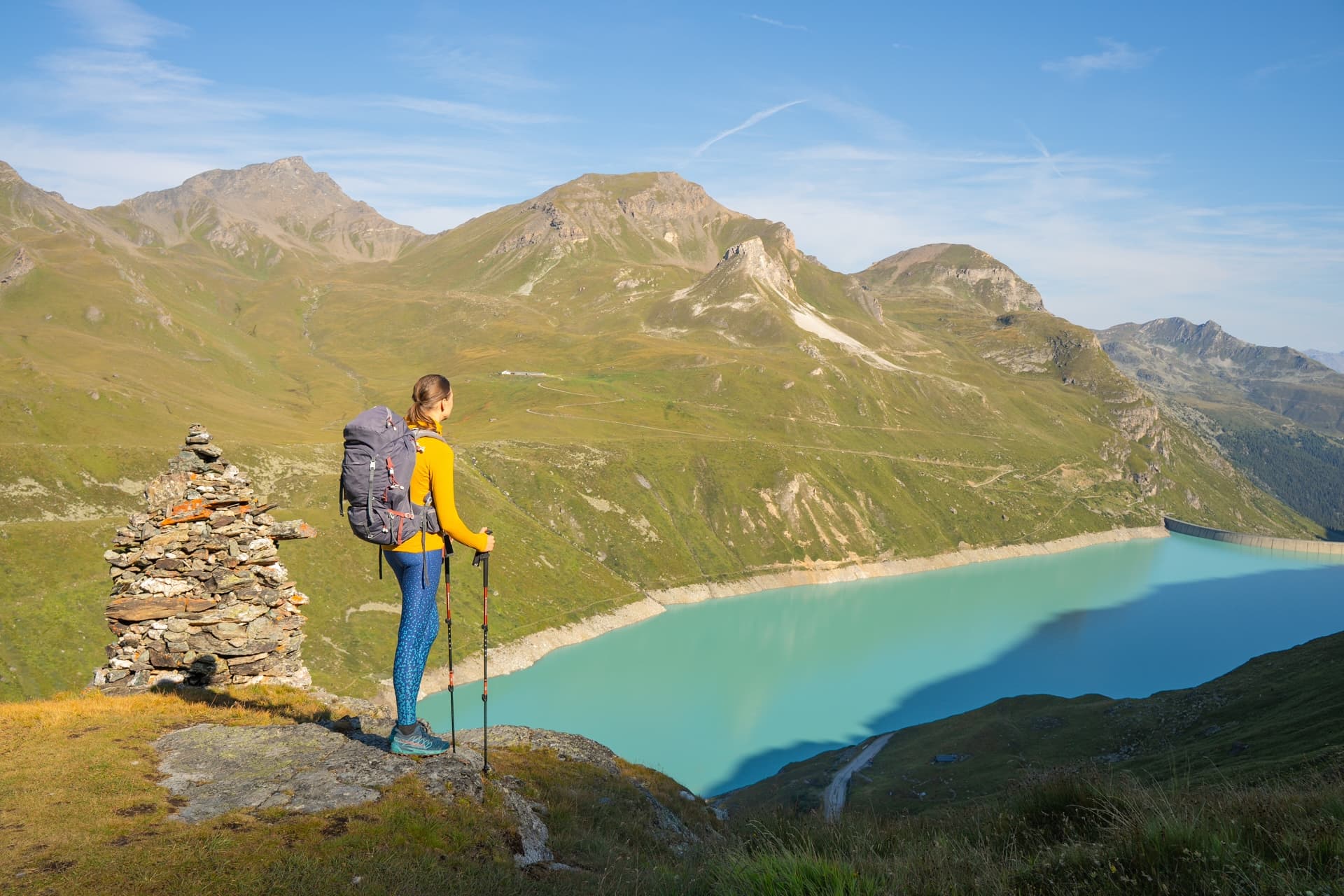 Hiker with backpack and poles overlooking turquoise Lac de Moiry reservoir and green mountains.