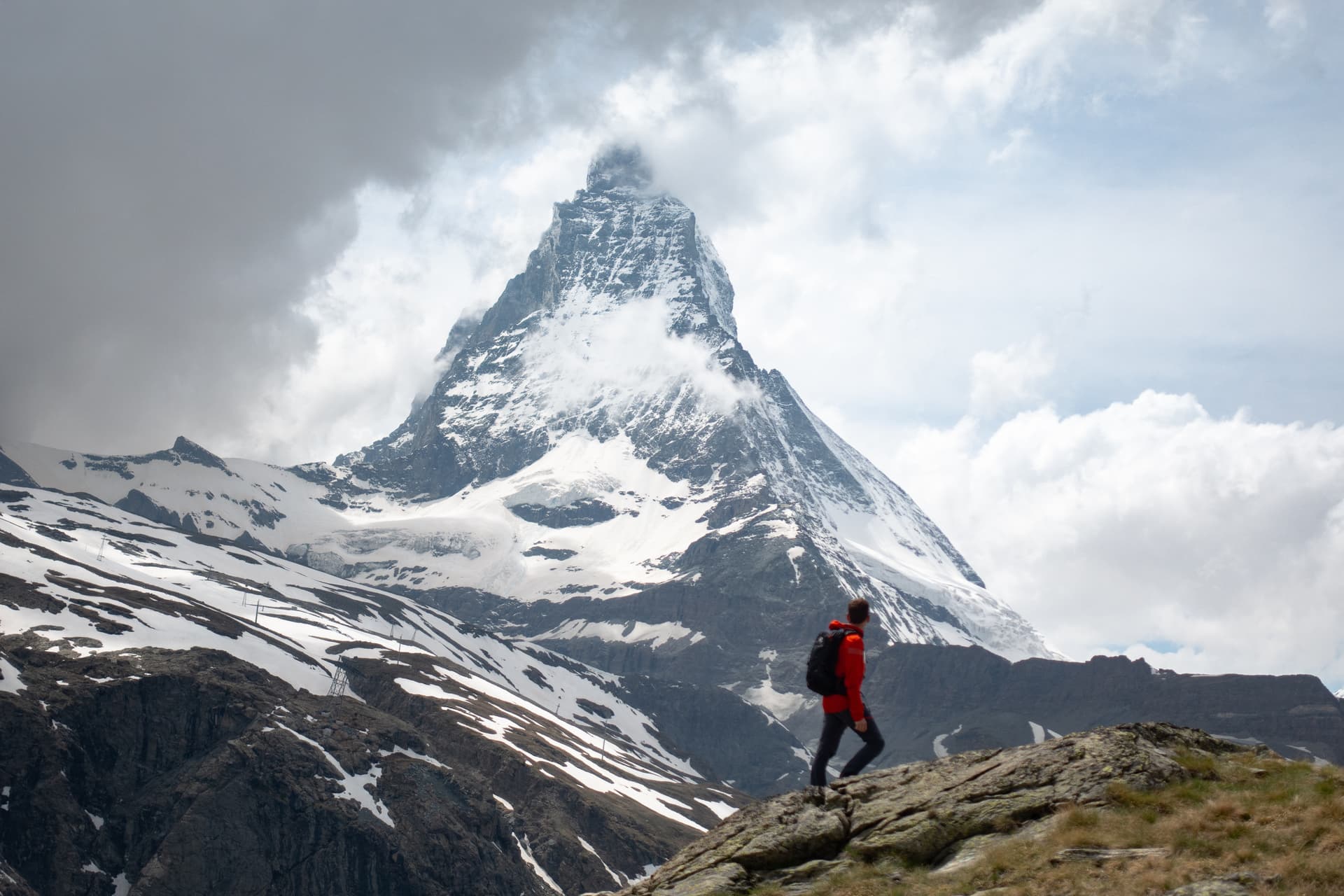 Hiker in red jacket walking near snow-capped Matterhorn mountain under cloudy sky