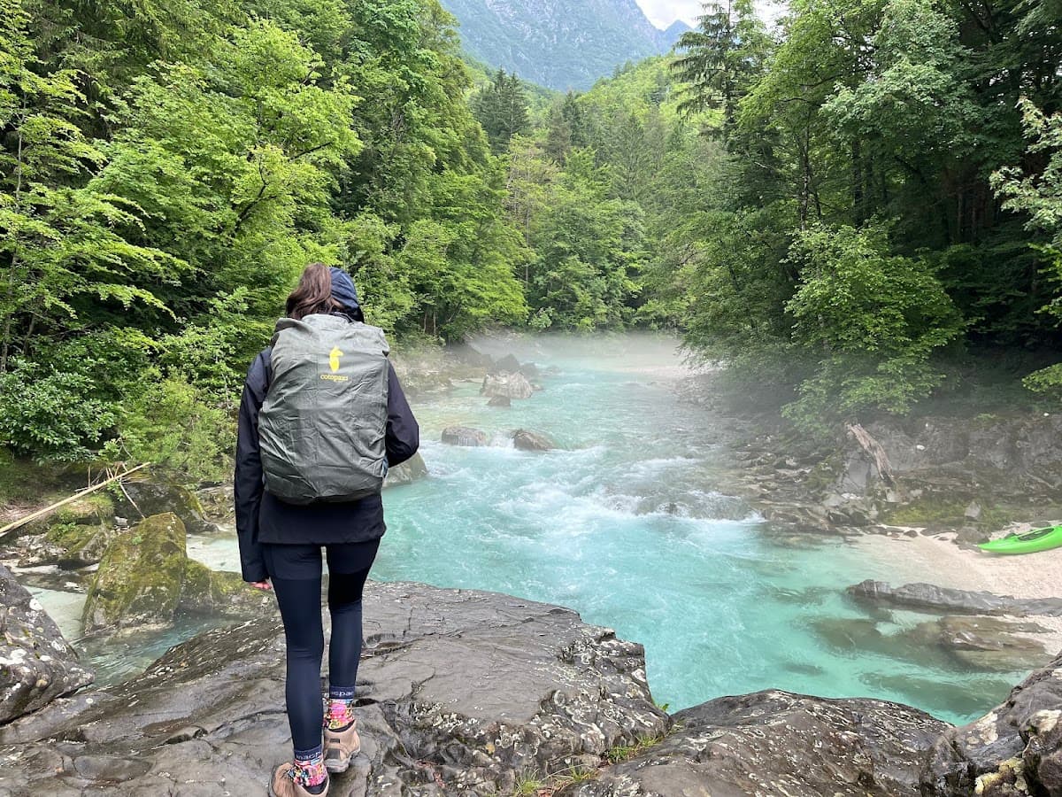 Hiker with backpack overlooks turquoise river rapids surrounded by lush green forest and mountains.