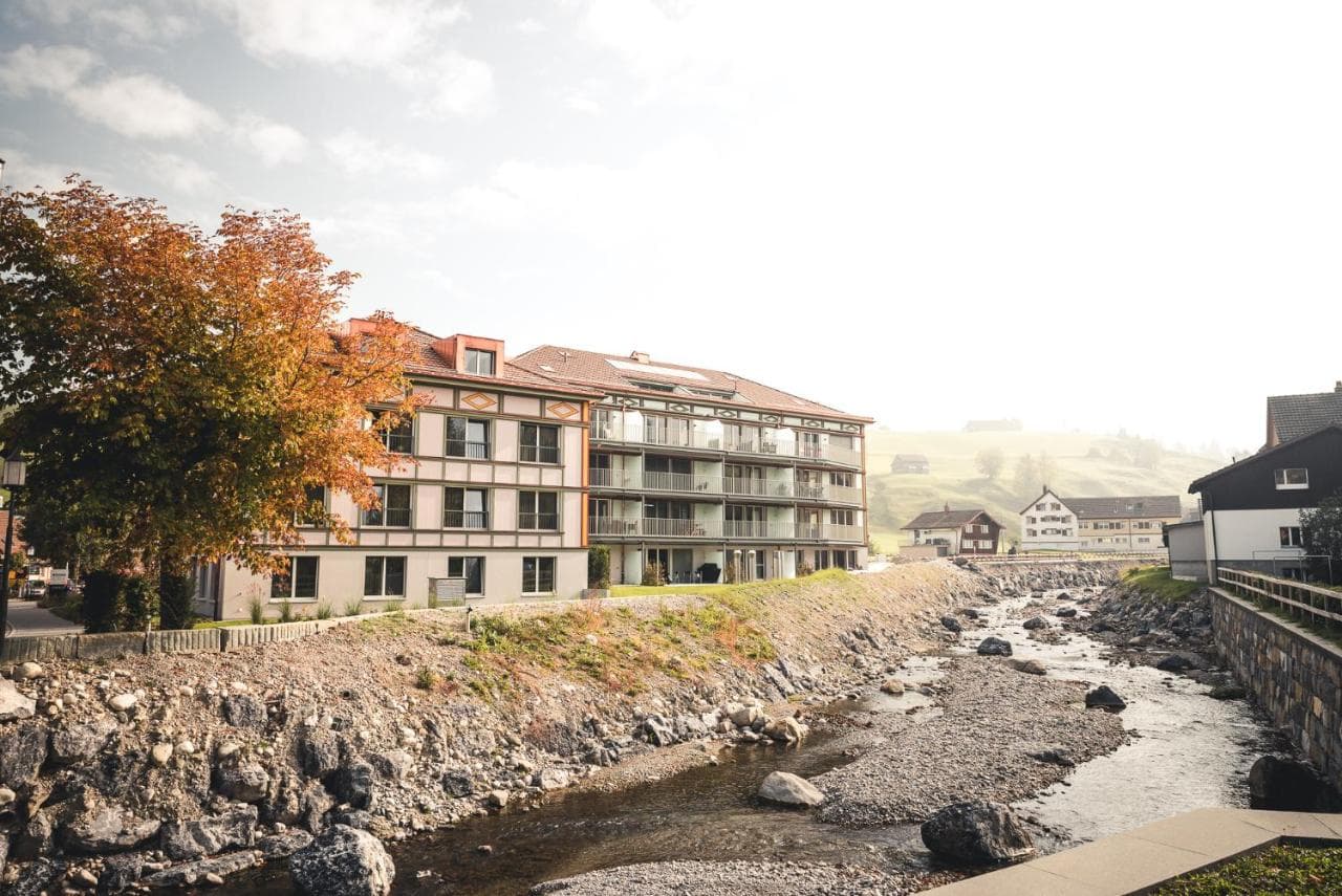 Modern lodge building next to a rocky stream with autumn foliage in a misty alpine village setting.
