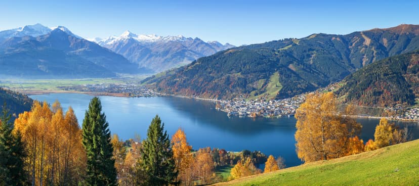 Panoramic view of beautiful autumn scene in the Alps with crystal clear Zeller lake, golden leaves and famous Kitzsteinhorn on a sunny day with blue sky in fall, Zell am See, Salzburger Land, Austria