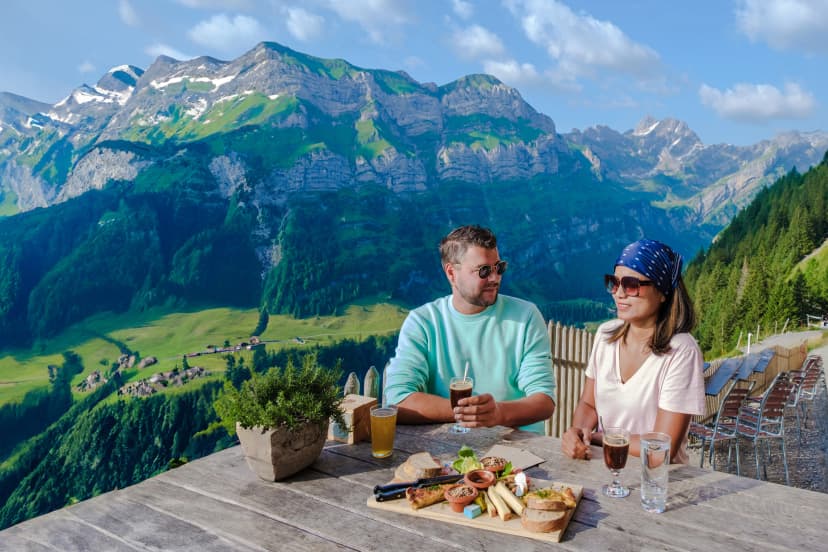 A couple of men and women drinking coffee at Berggasthaus Aescher in den Appenzeller Alpen at sunset, a restaurant under a cliff at the mountain Ebenalp in Switzerland, Appenzell