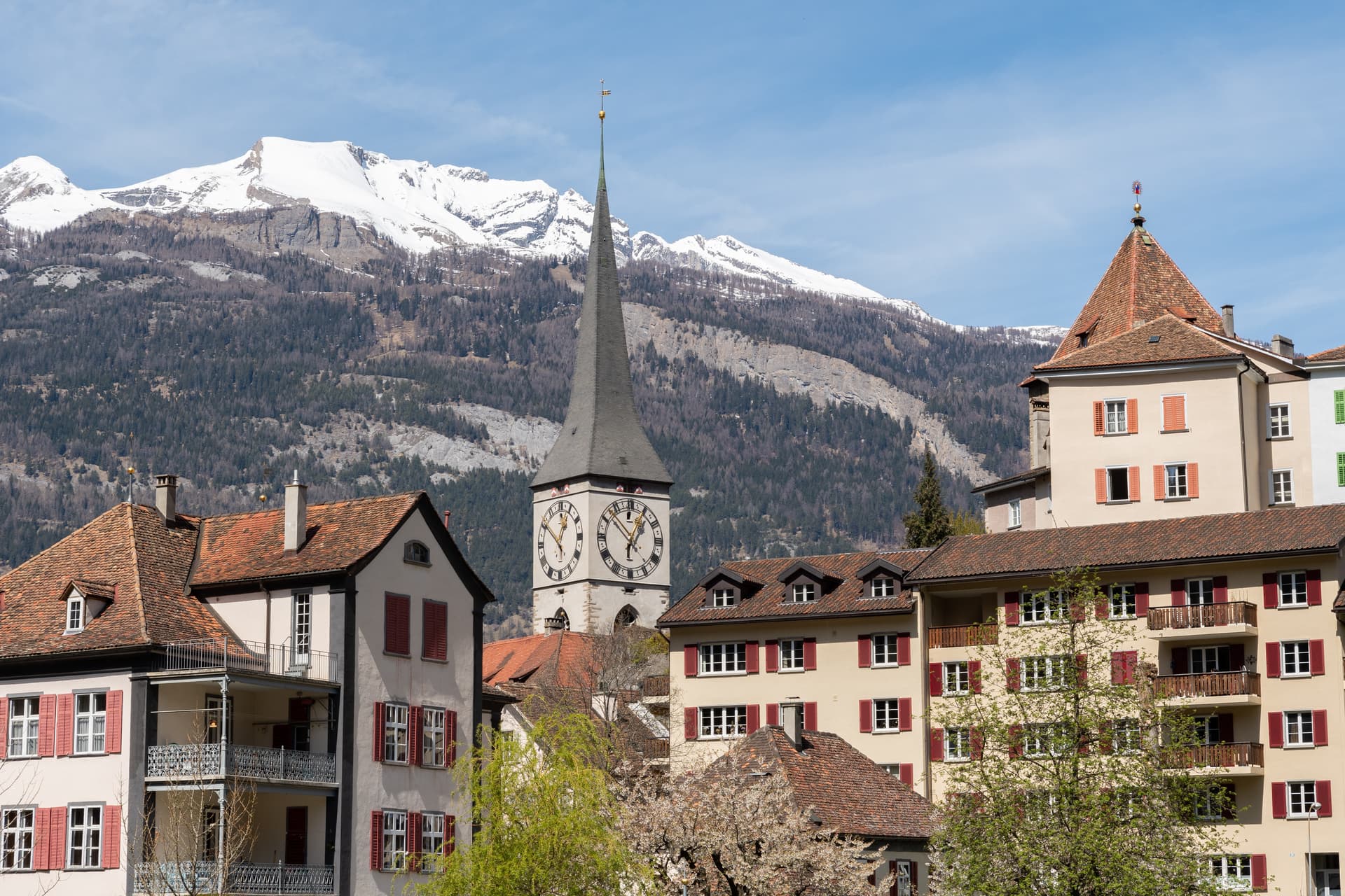 Town buildings with red shutters and a clock tower against snow-capped mountains.