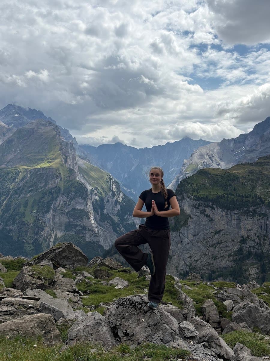 Person doing yoga pose on rocky mountain overlook with alpine peaks and cloudy sky