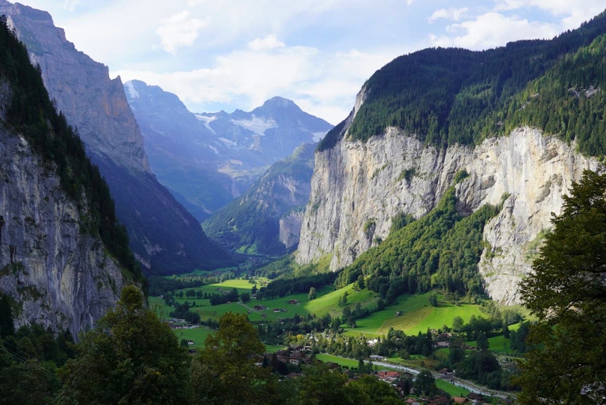 Alpine valley with steep rock cliffs, green fields, and distant snowy mountain peaks