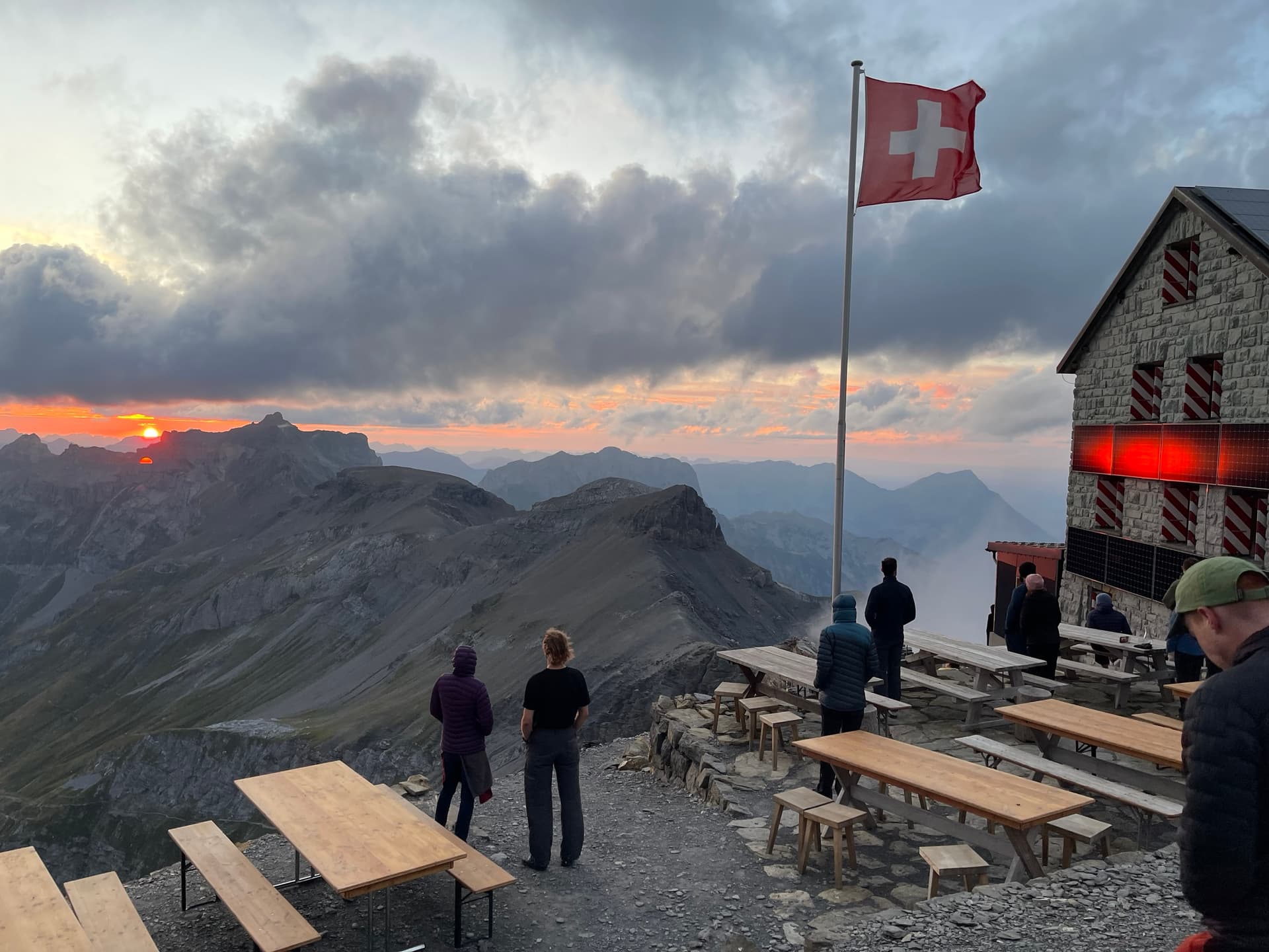 People standing on mountain hut terrace at sunset with Swiss flag and alpine views