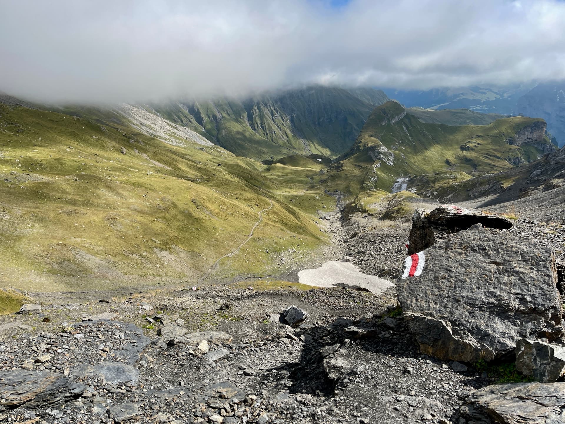 Red and white trail marker painted on rock overlooking alpine valley with mountains and clouds