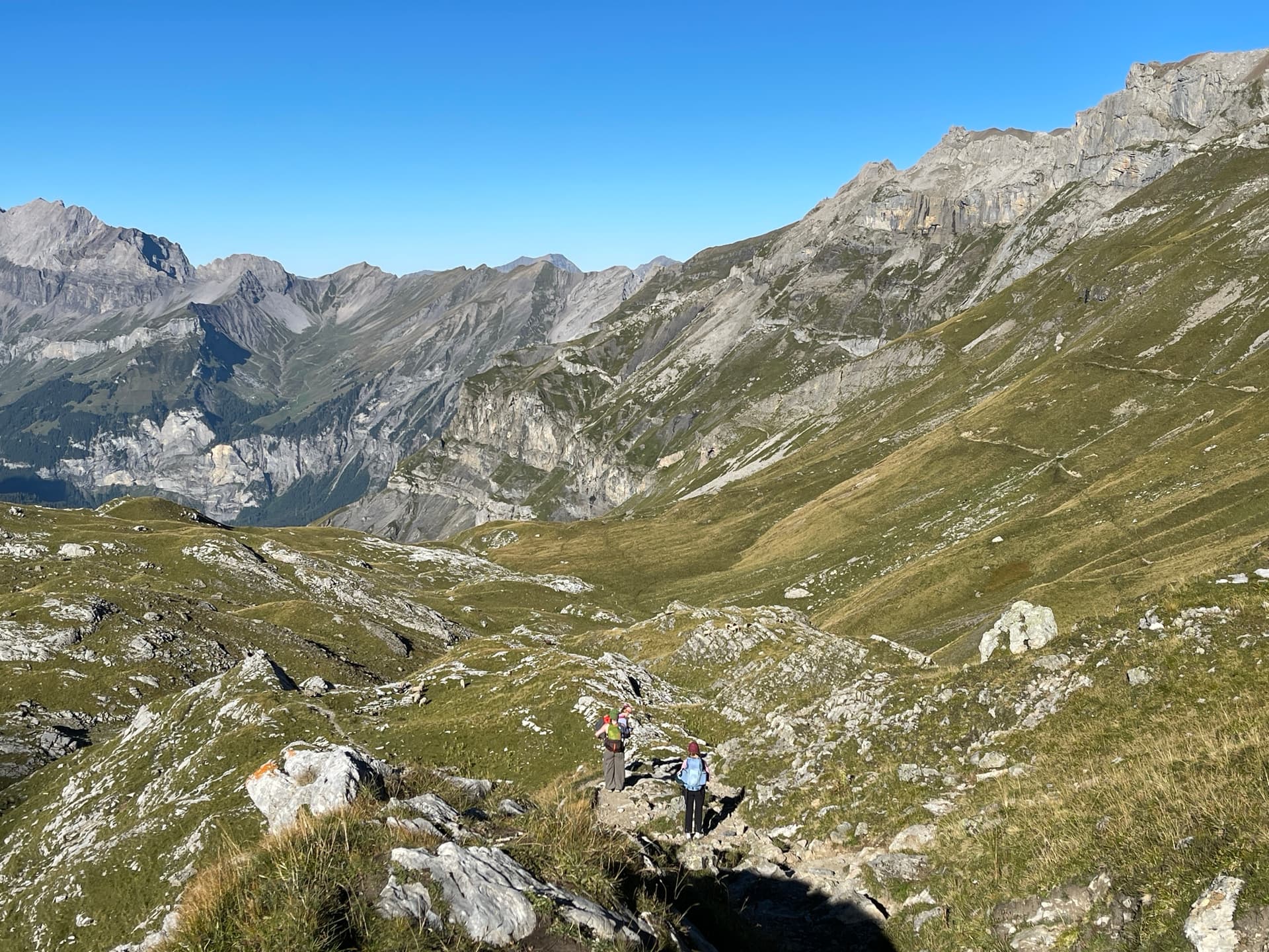 Two hikers on a trail in a wide alpine mountain valley with green slopes and rocky terrain under a blue sky.