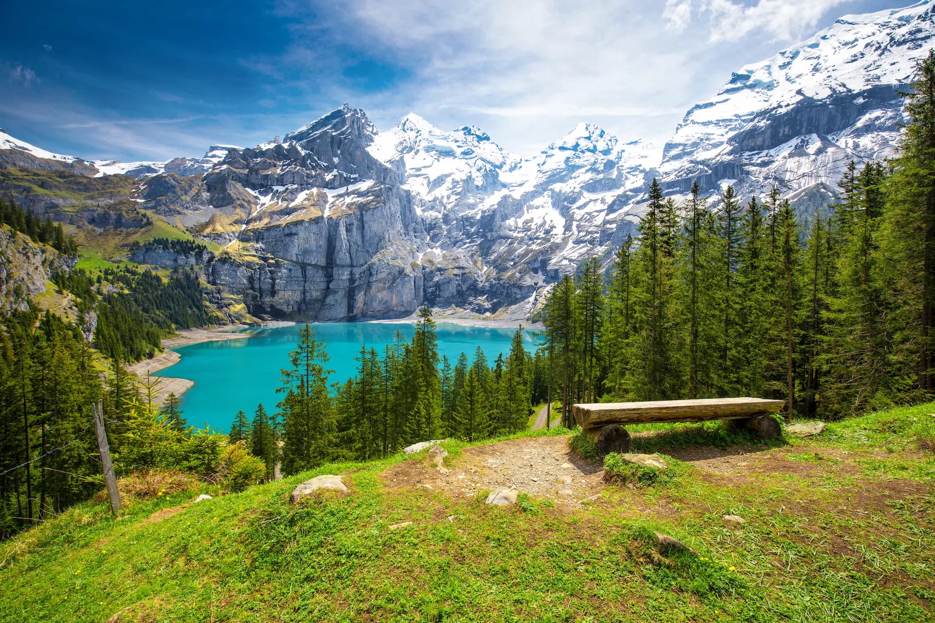 Turquoise Oeschinnensee lake below snow-capped Swiss Alps mountains with pine forest and wooden bench.