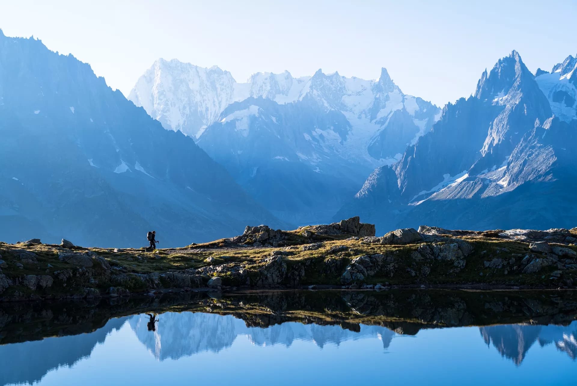 Hiker by alpine lake reflecting snowy Chamonix mountains in bright morning light.