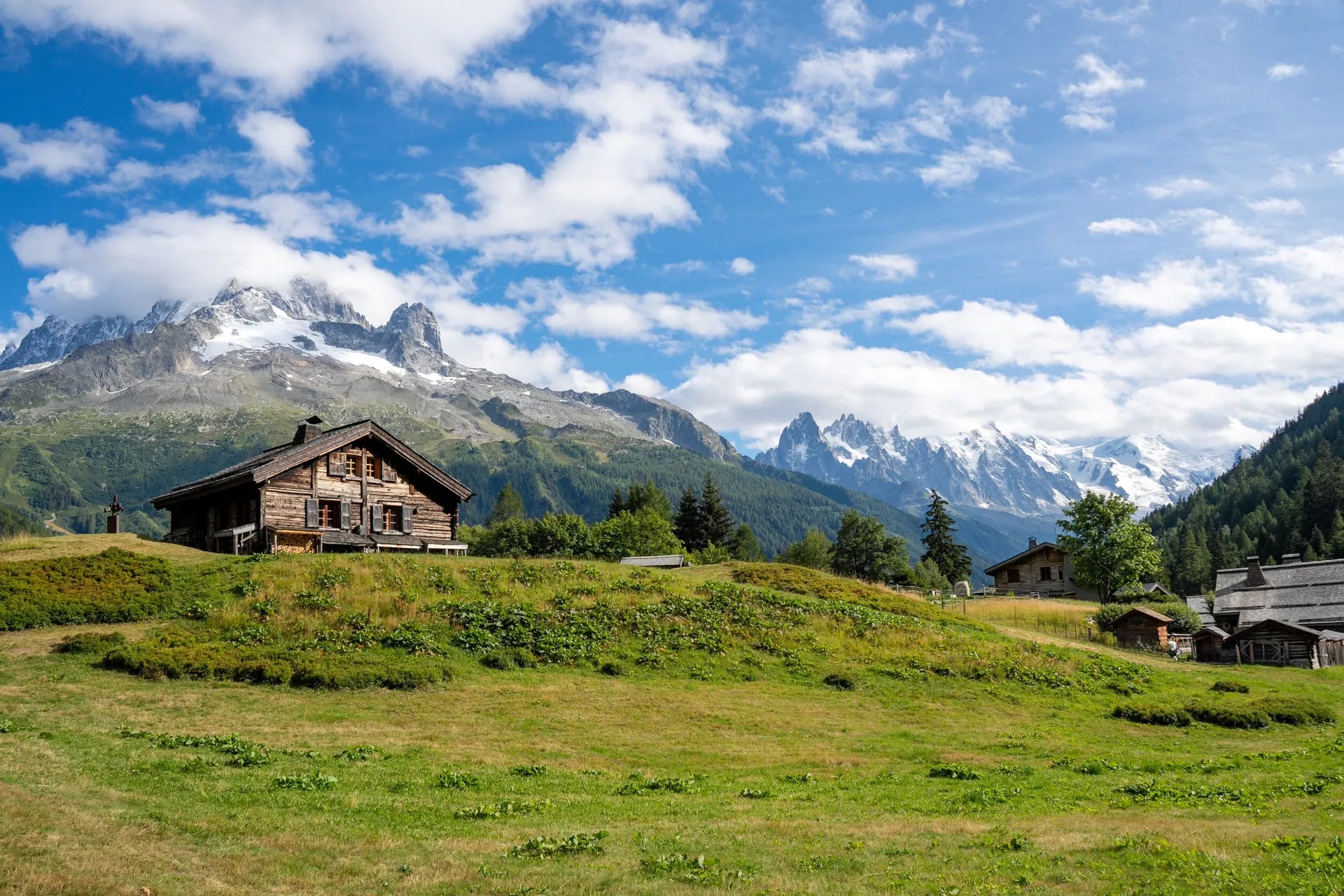 Wooden alpine chalets on a grassy hill with snow-capped mountains under a blue, cloudy sky.