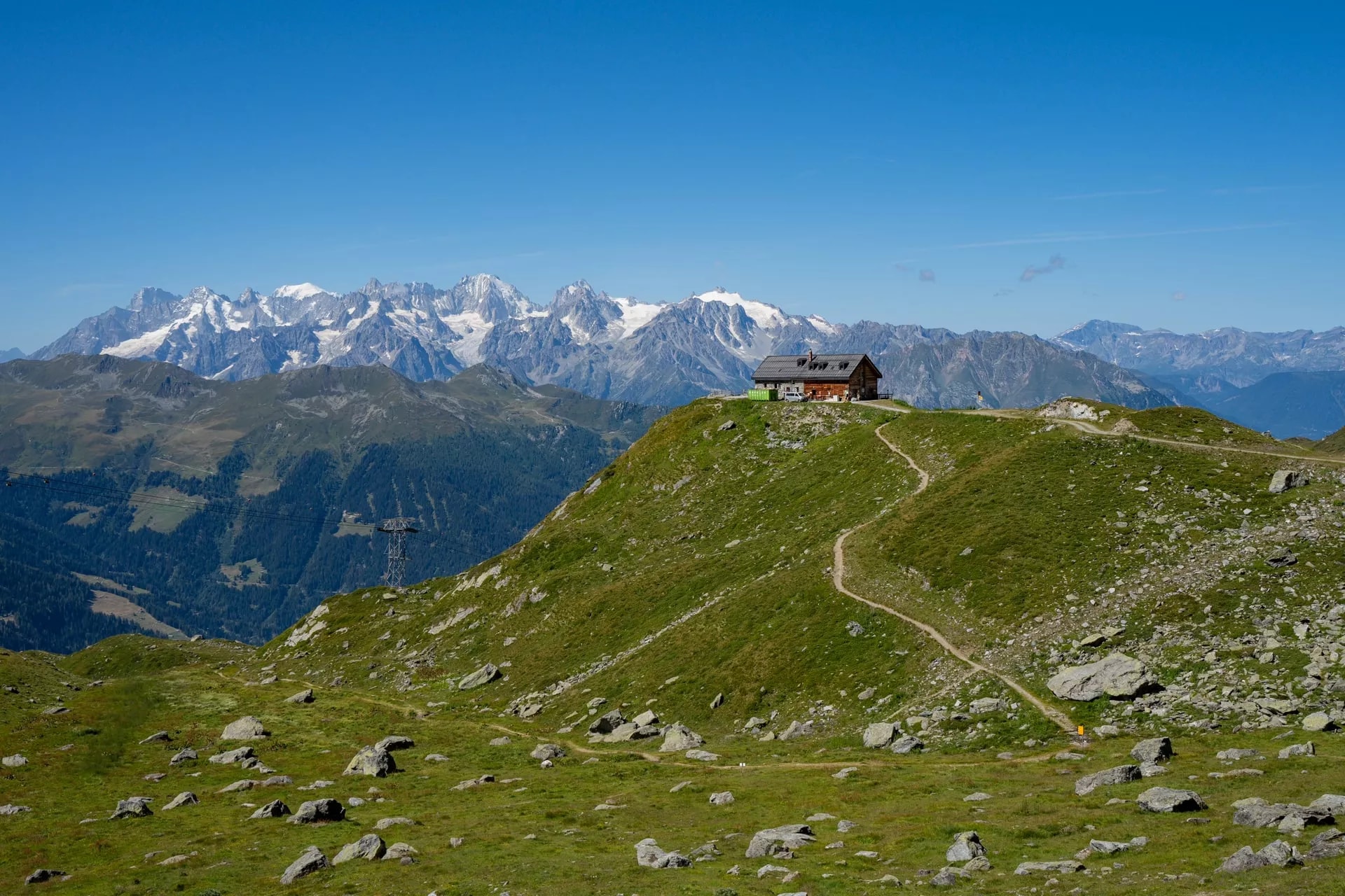 Mountain hut on grassy peak with hiking path, snowy peaks in background under blue sky