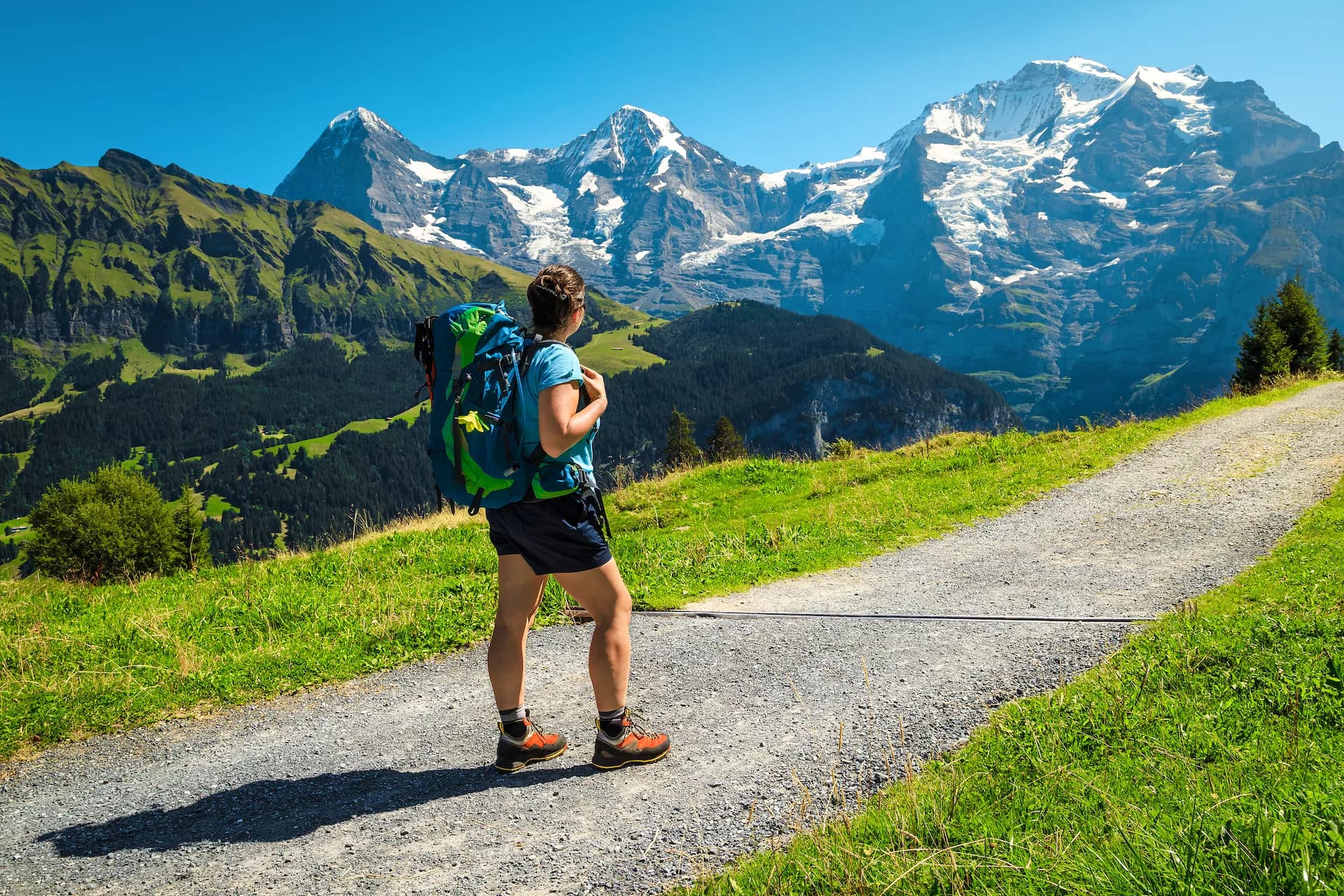 Hiker with backpack enjoying view of snow-capped mountains above Mürren on sunny day.