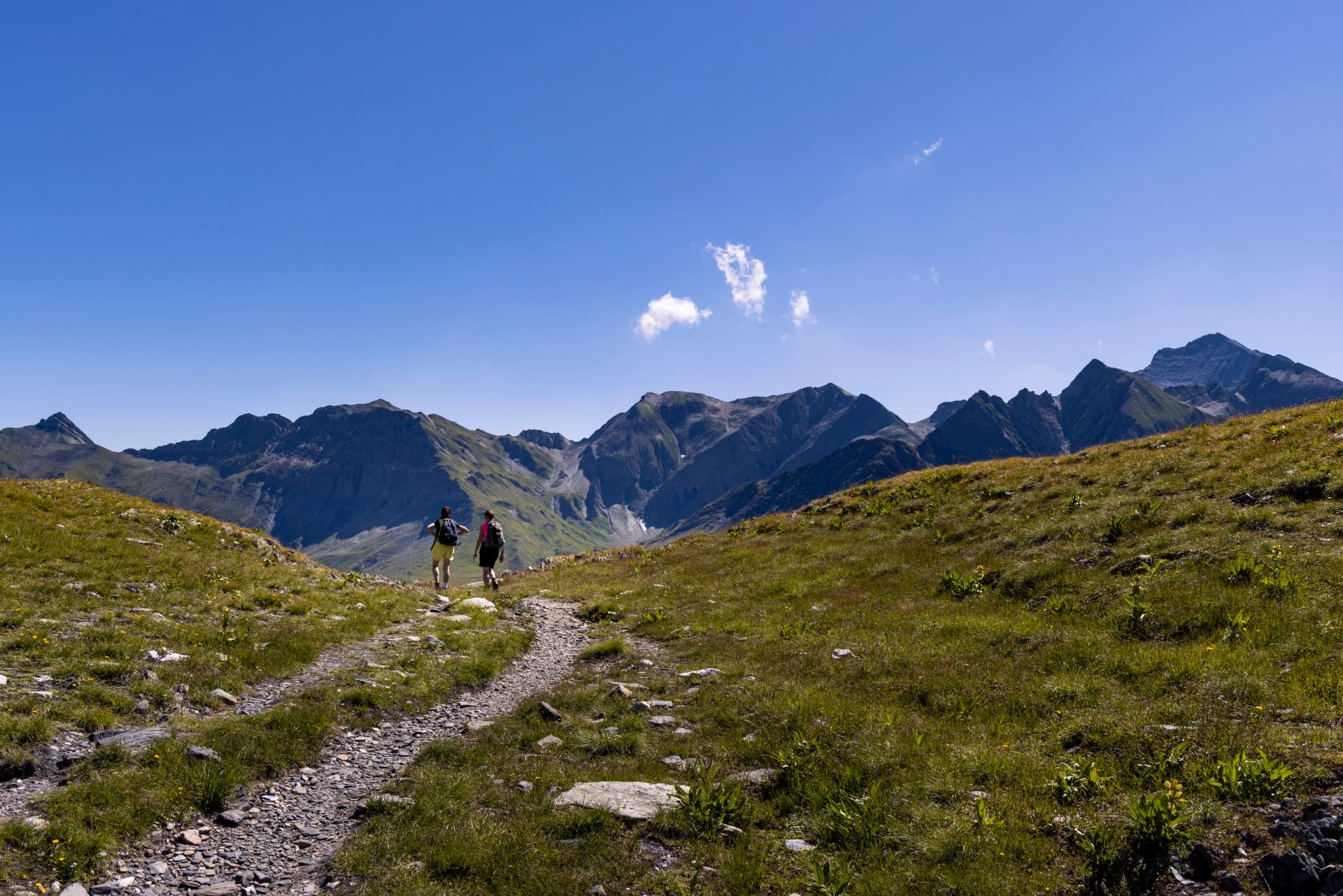 Two women walk on a winding alpine trail between mountains in late summer on a sunny day in the Greina Plateau, in Switzerland.